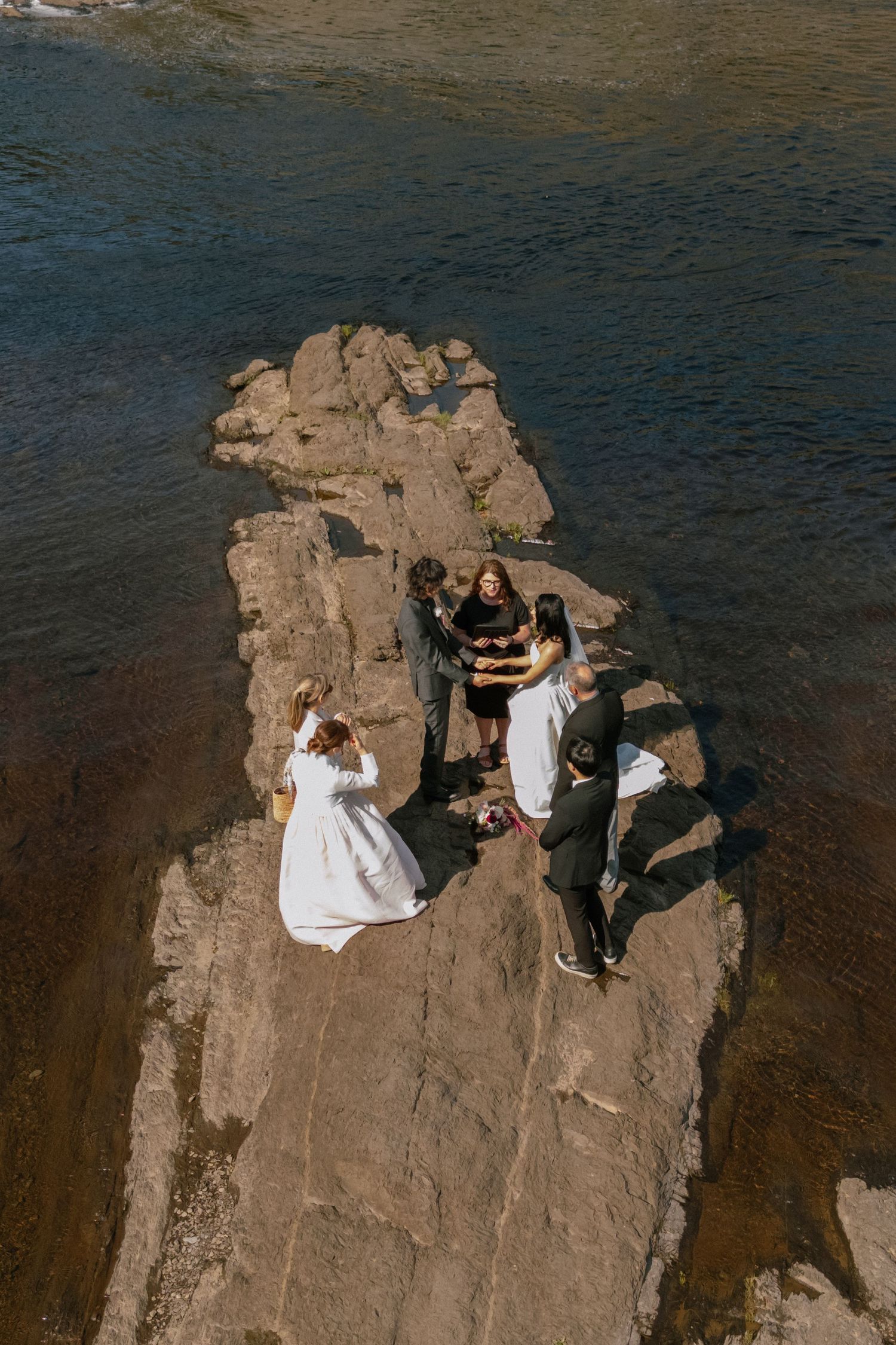 A wedding couple explores near a majestic waterfall on rocky terrain surrounded by lush forest.