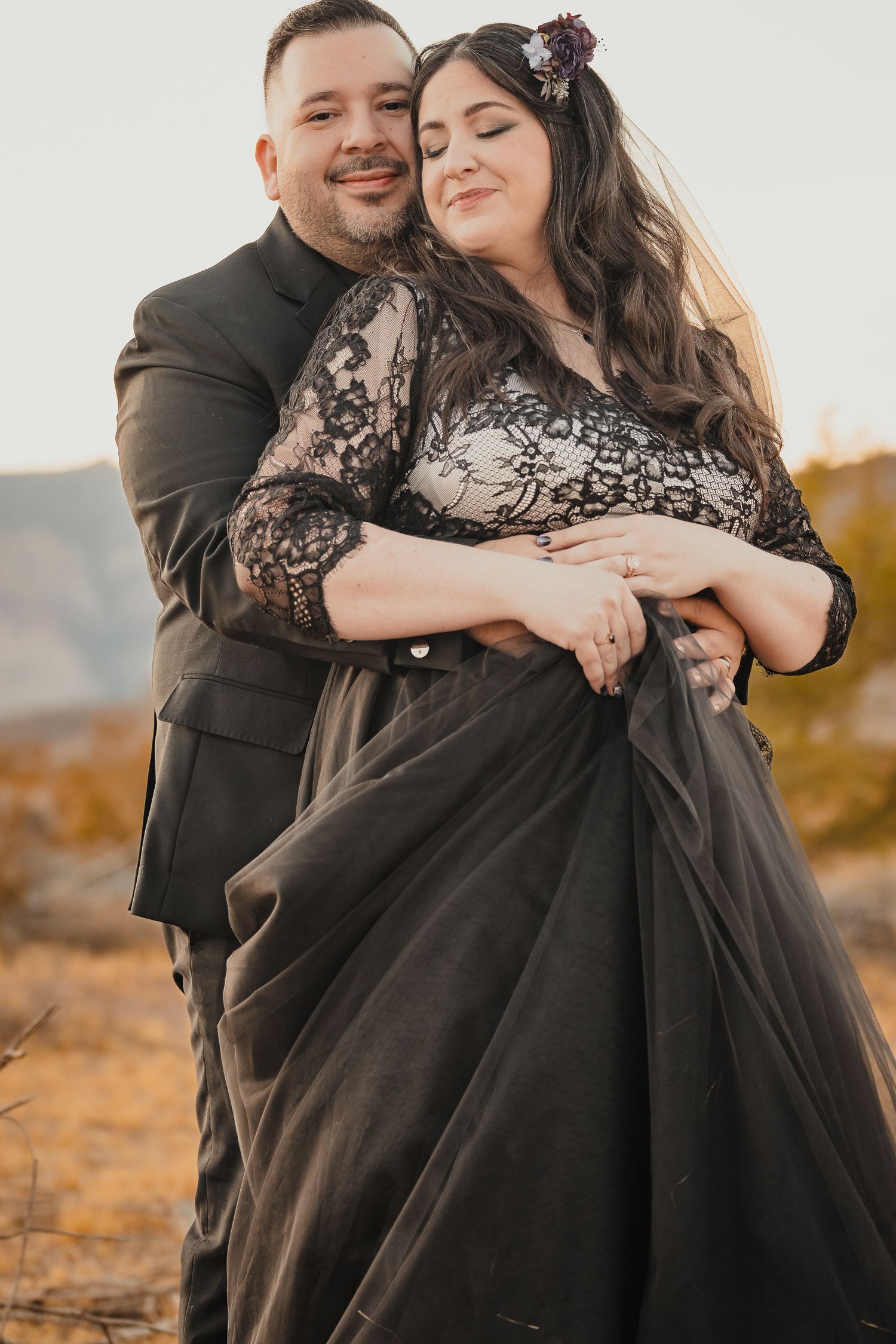 A romantic outdoor desert portrait features a couple embracing, with the subject wearing an elegant black lace wedding dress.