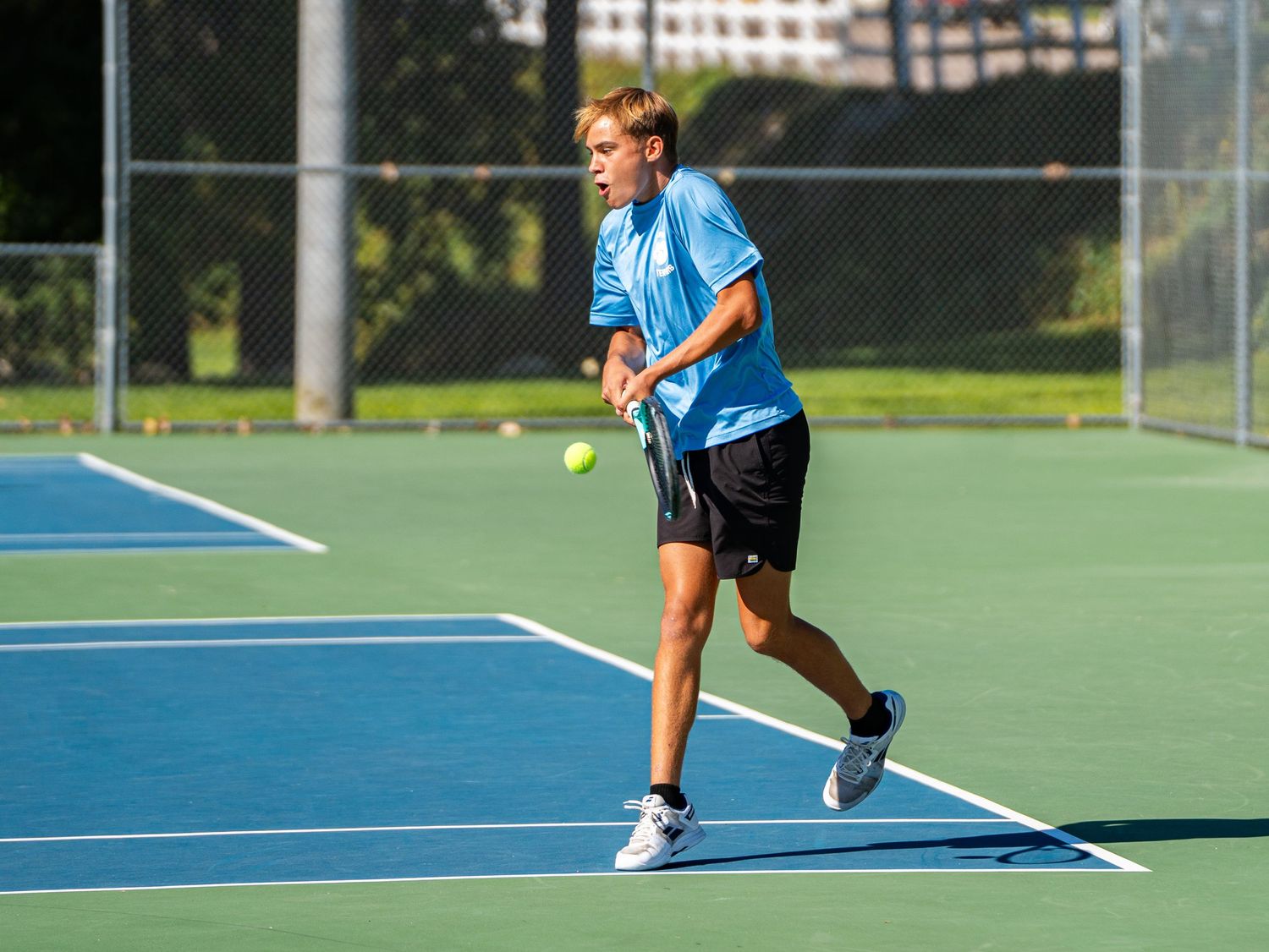 Tennis player in light blue shirt practices form on outdoor court.