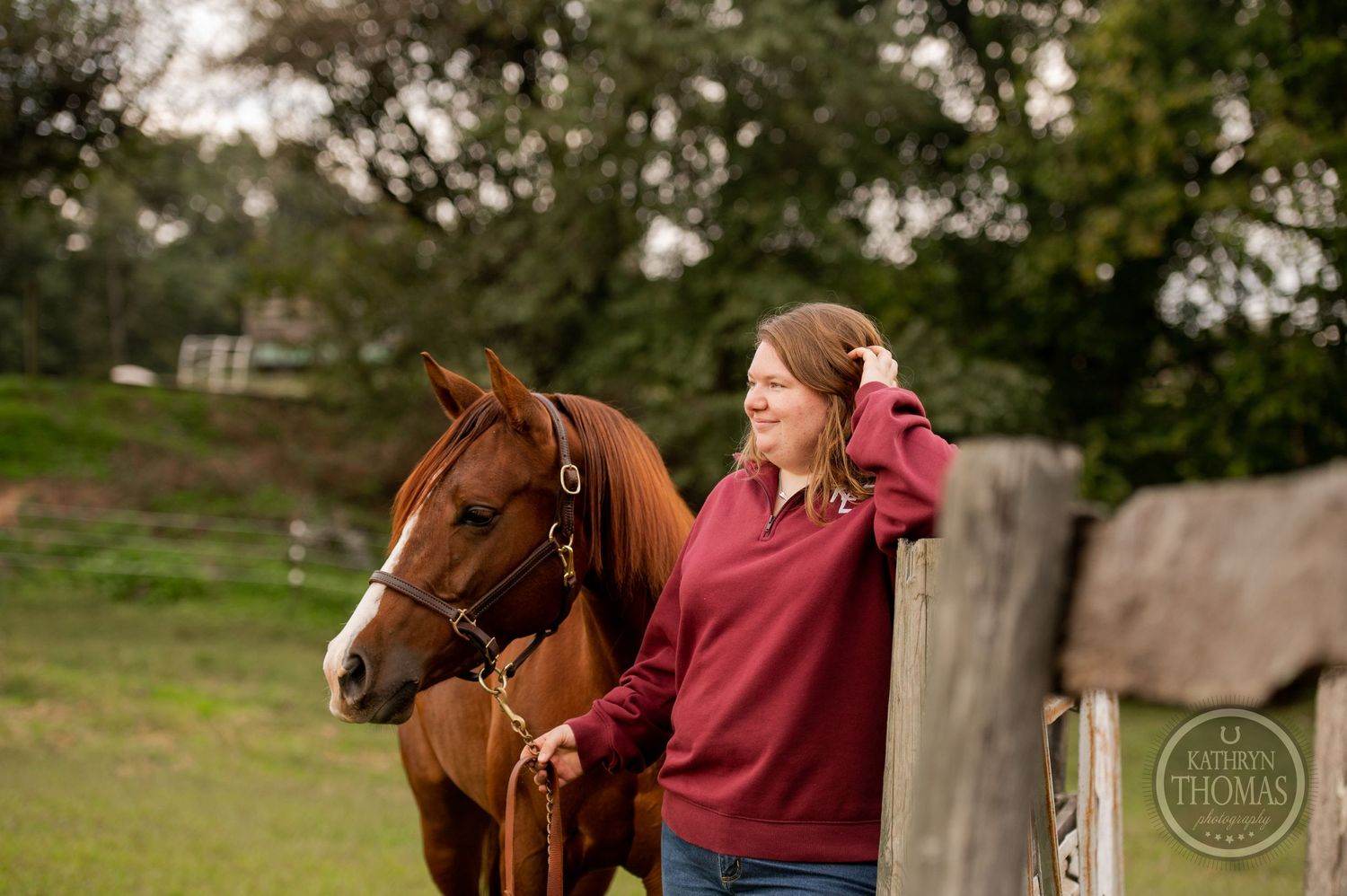A chestnut horse stands beside someone in a burgundy sweater against a pastoral background.