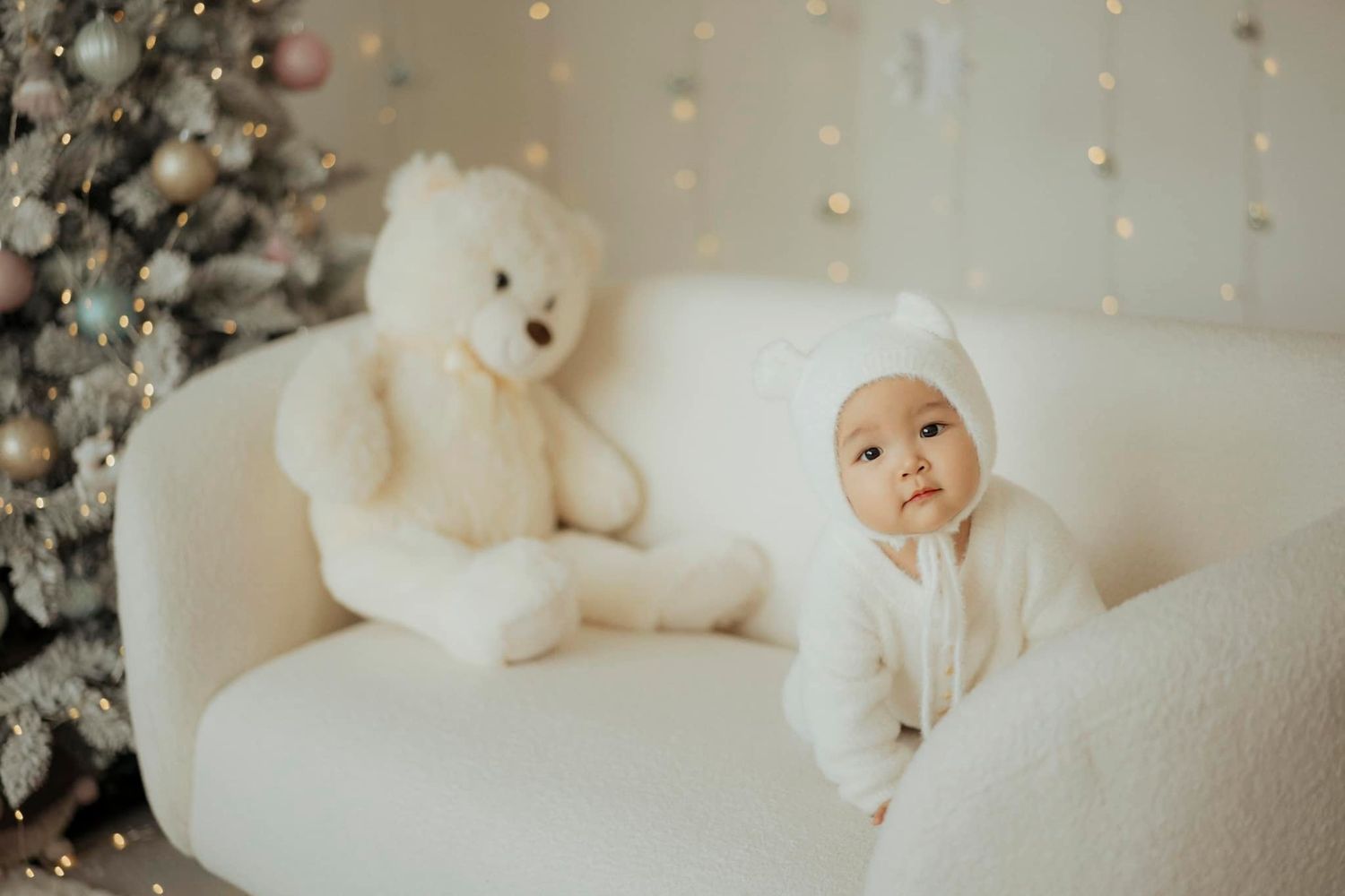 A small baby in white sits on a cream couch next to a plush teddy bear near holiday decorations.