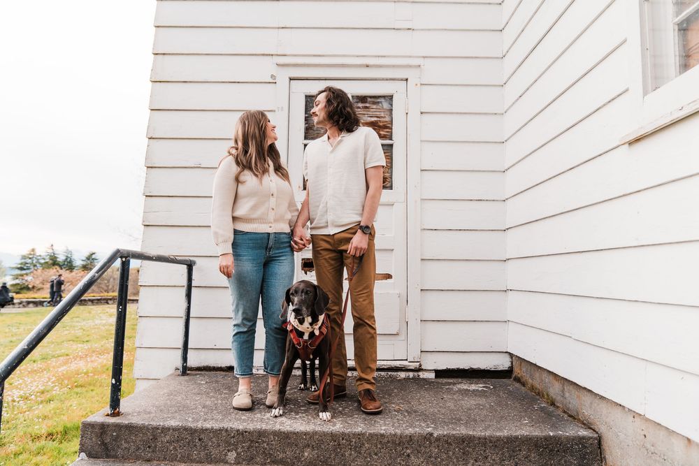 A couple and their black dog stand on steps outside a white building, smiling at each other.