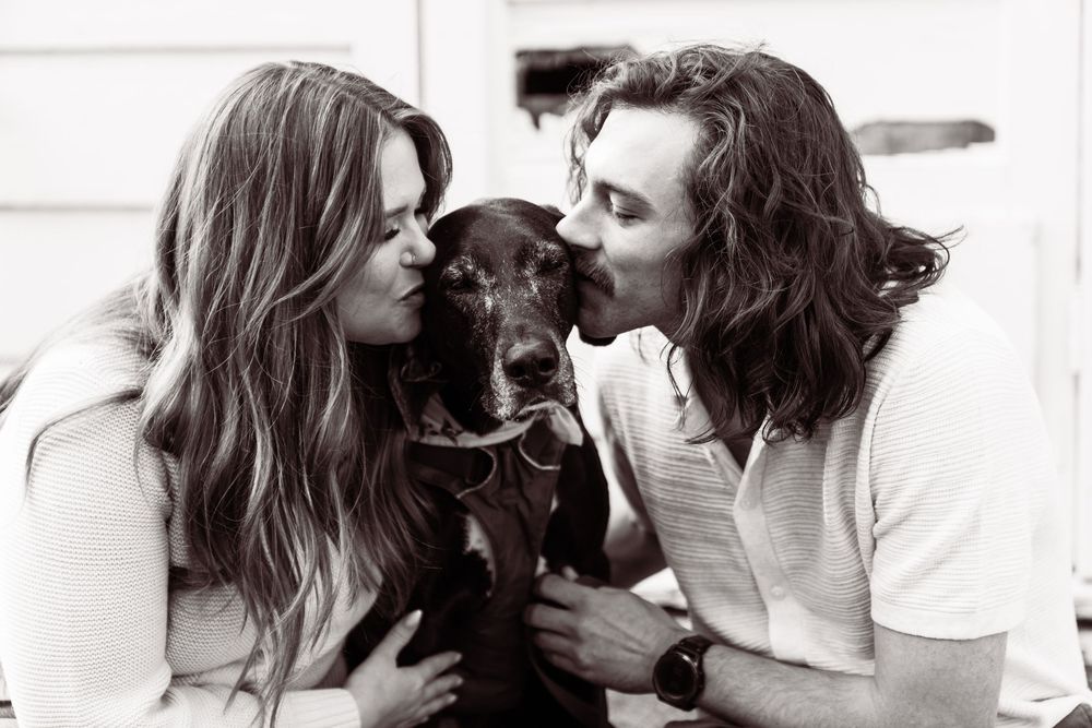 A black and white photo of a couple kissing their Labrador dog on both sides.