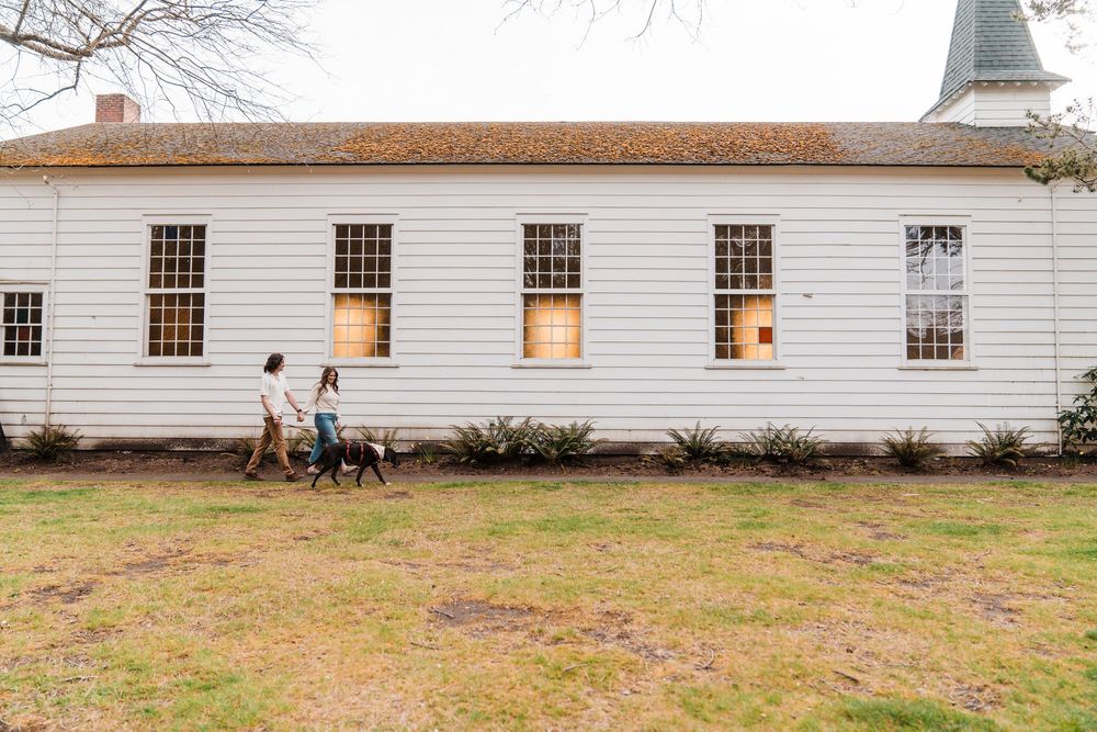 A couple walks their dog past a large white historic building on a grassy lawn.