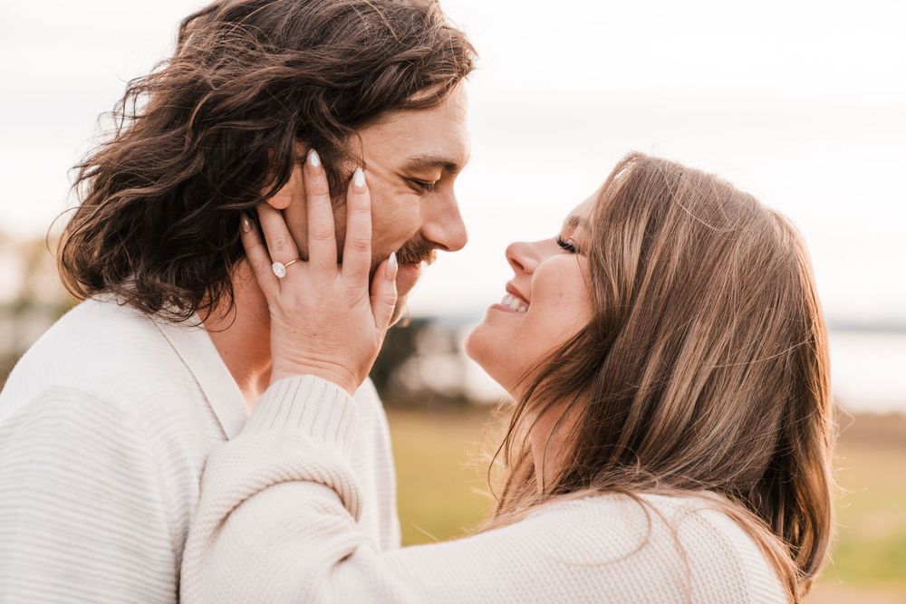 Couple sharing a romantic moment, smiling at each other, woman wearing engagement ring, outdoors with soft natural light.