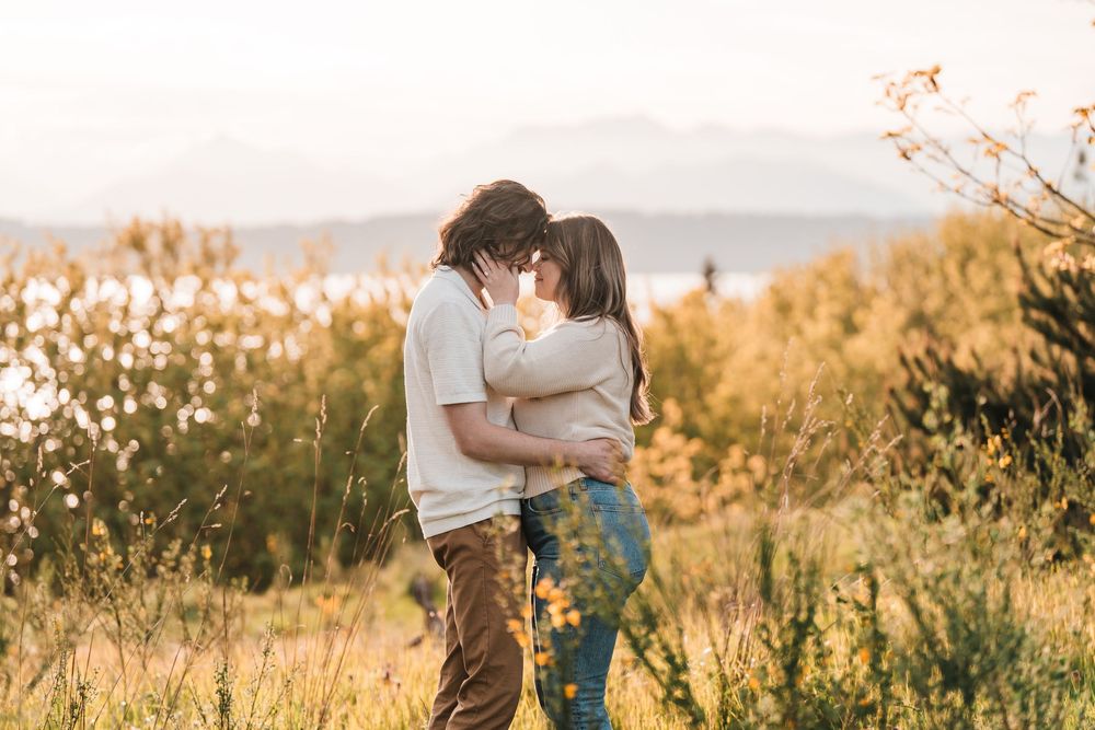 Couple sharing a kiss in a golden field with a serene lake and mountains in the background at sunset.