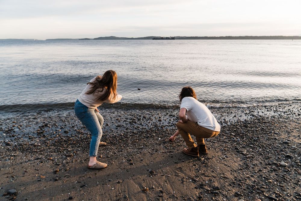 Couple crouching together on a rocky beach shoreline, exploring stones near calm water at dusk.