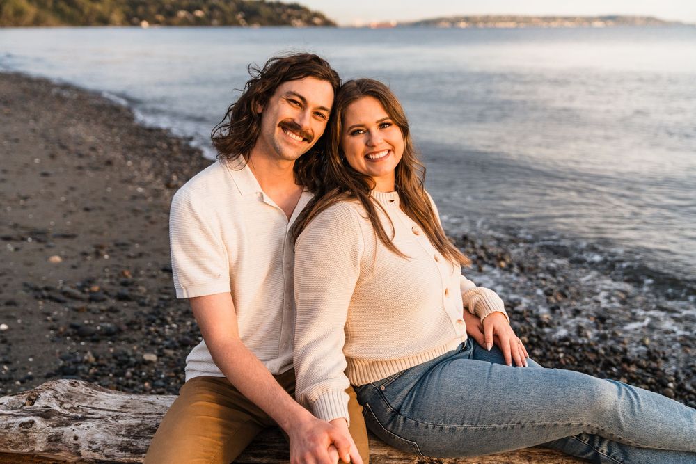 Smiling couple sitting on driftwood along a pebble beach at sunset, holding hands and posing together.