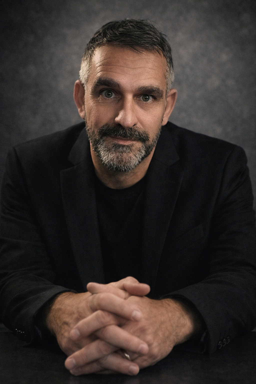 Professional portrait of a man in a black suit and tie against a textured gray background.
