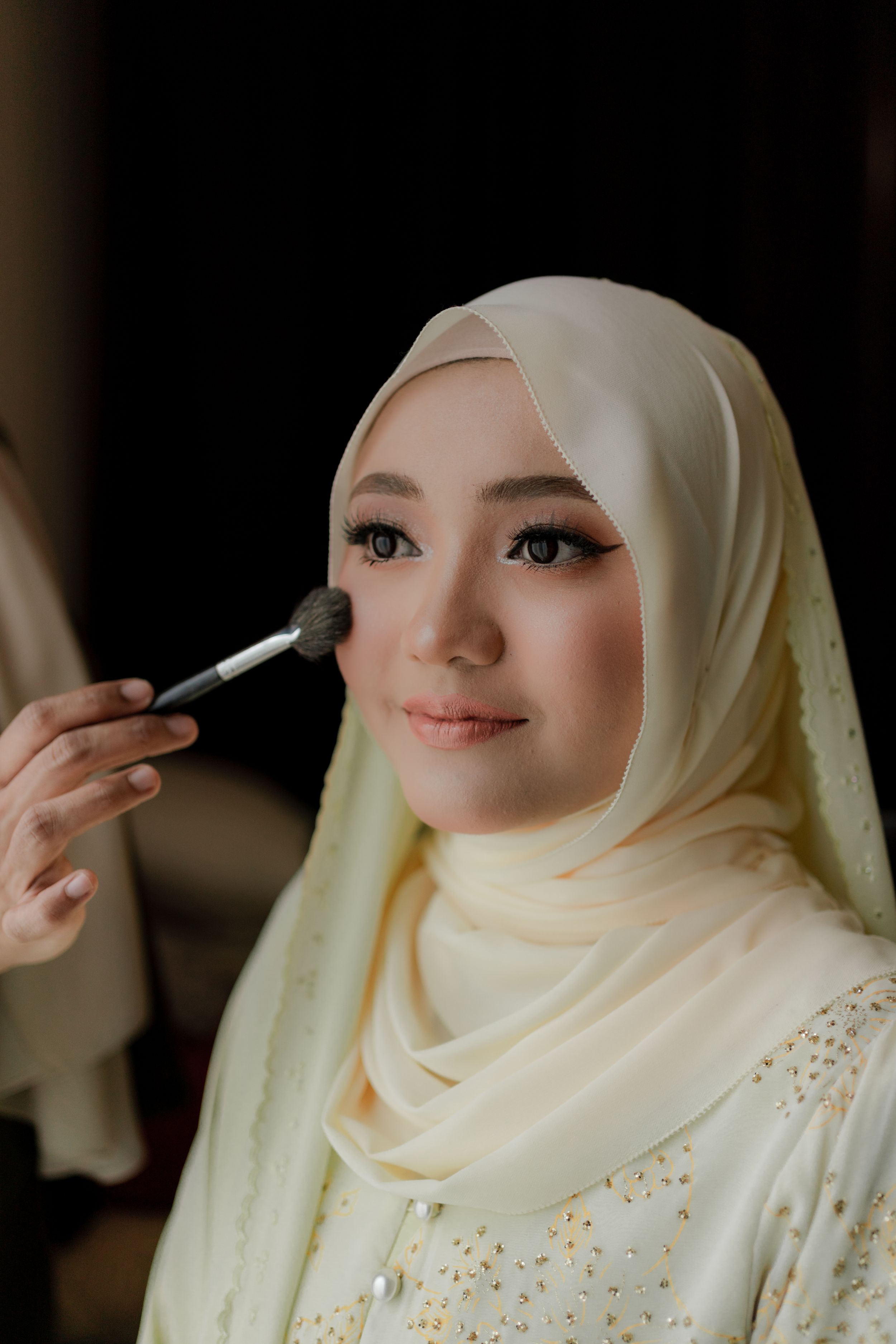 A makeup artist applies cosmetics to a bride wearing a light hijab and embellished dress.