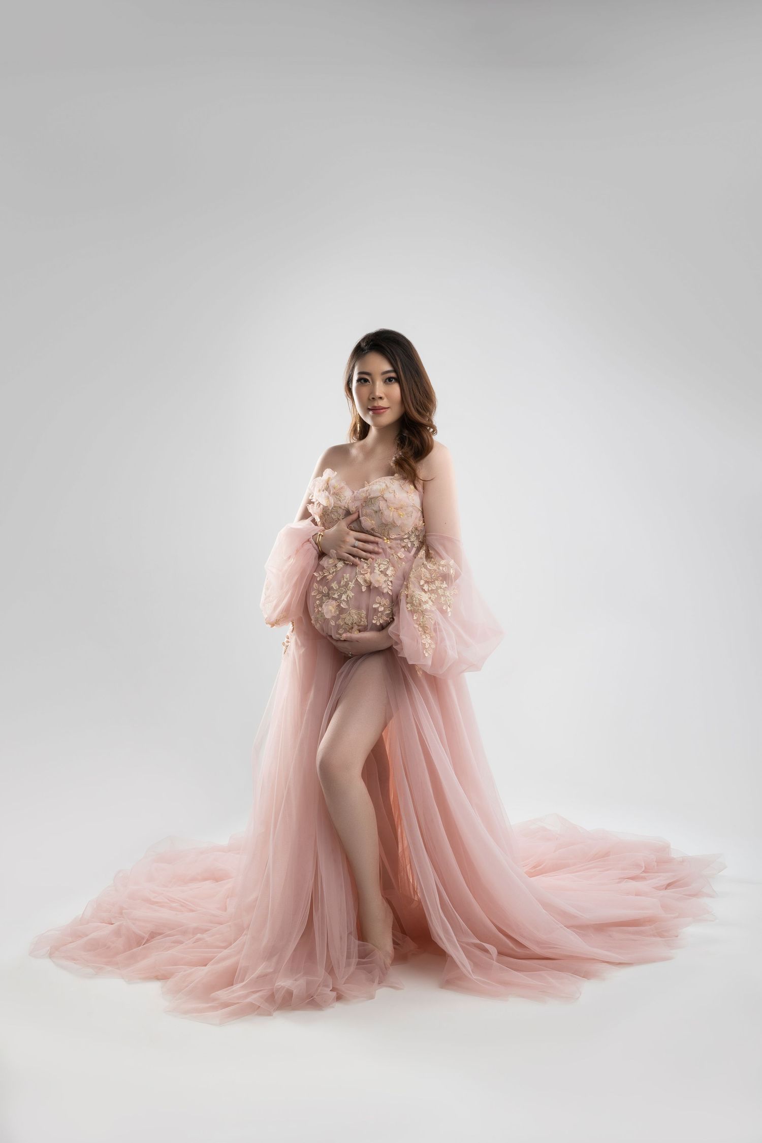 A pregnant woman poses in an elegant pink tulle gown with long sleeves and a high slit against a light gray backdrop.