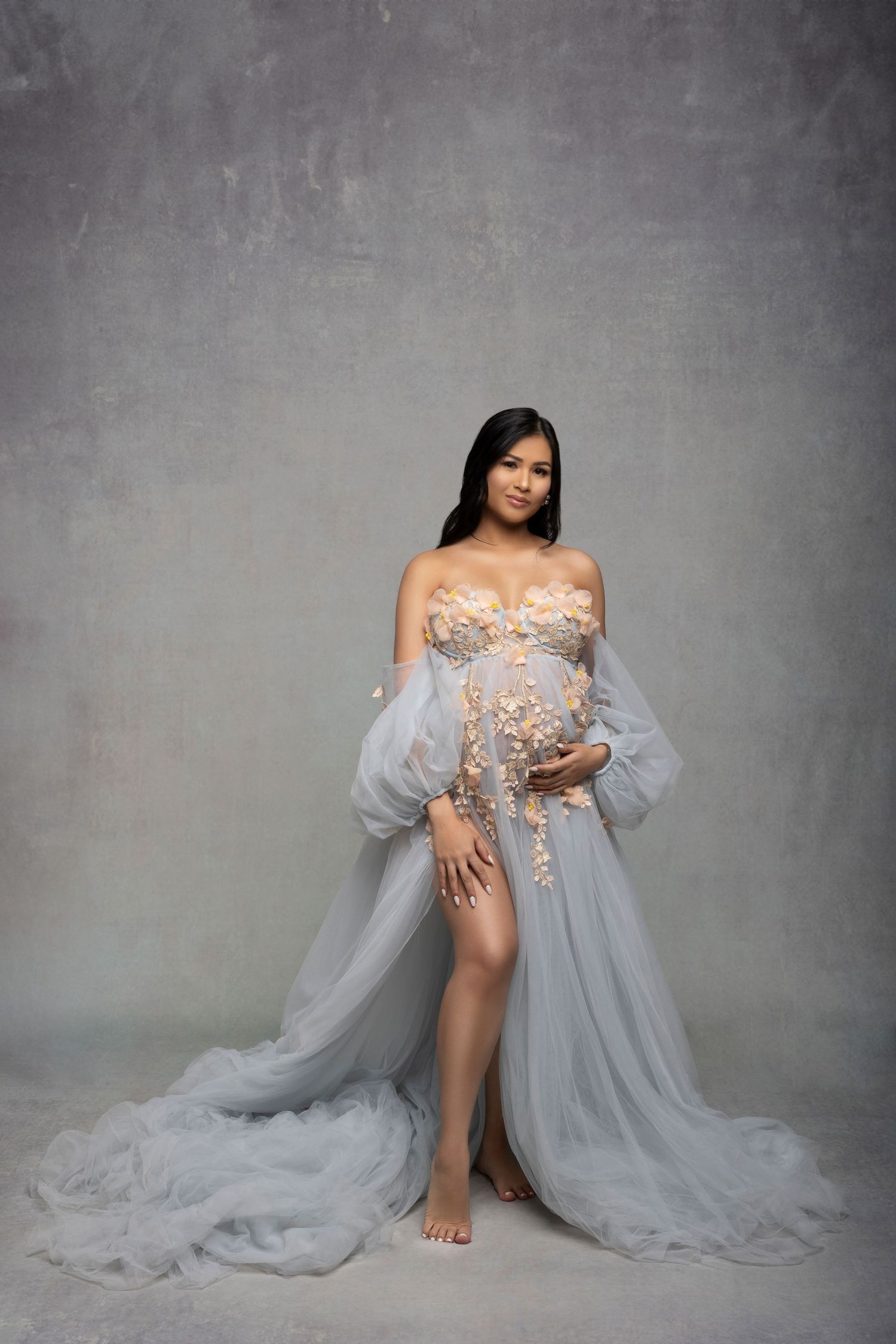 An elegant maternity photo featuring a flowing gray sheer gown with floral appliques against a textured gray backdrop.