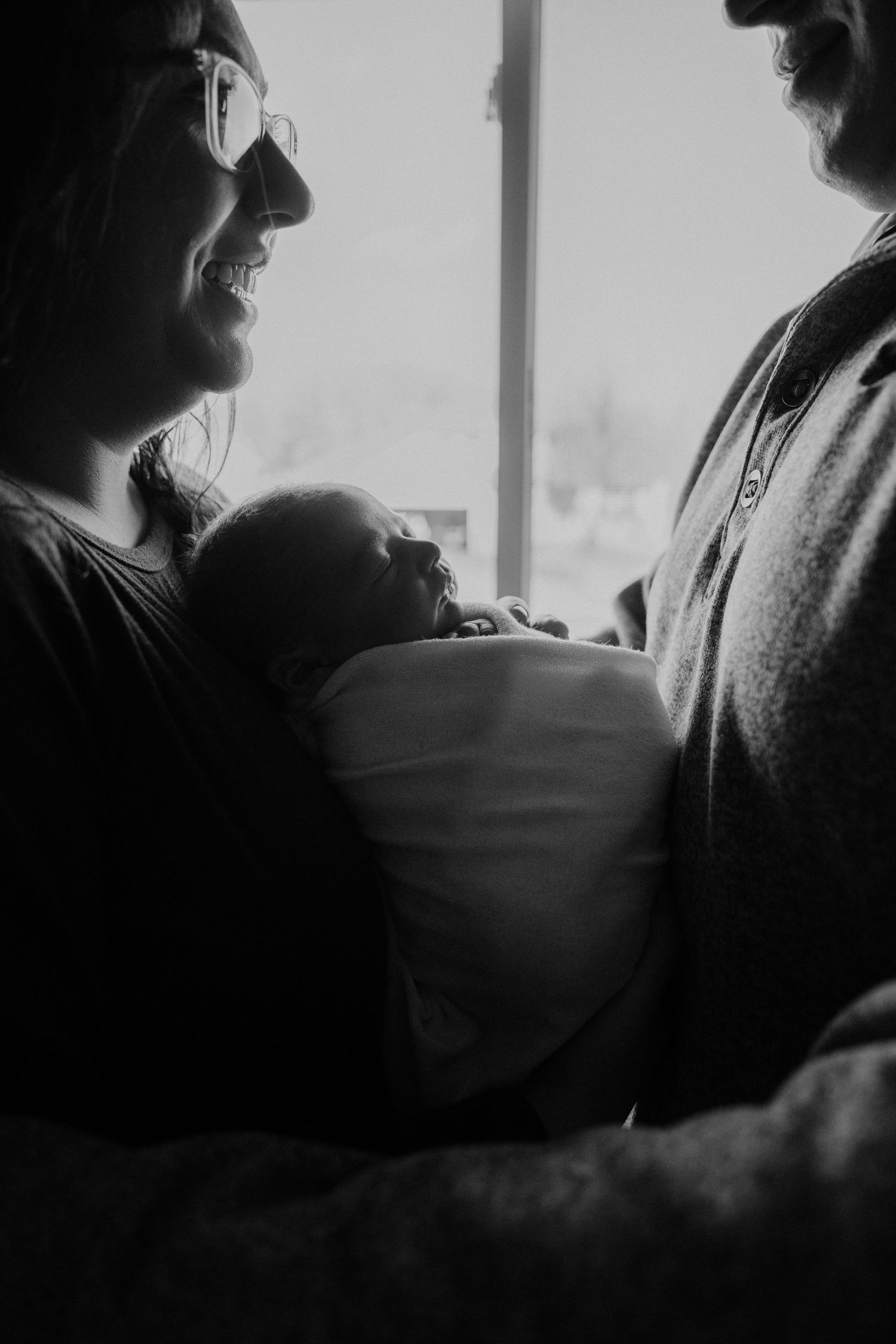 A tender black and white silhouette of parents holding their newborn baby near a window.