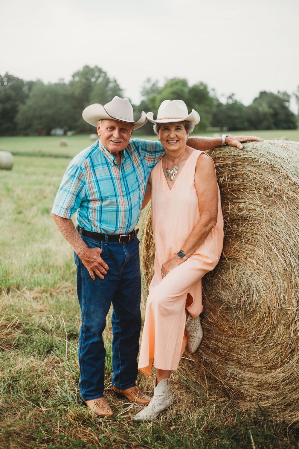 A country couple in western wear posing by a hay bale in a rural field.