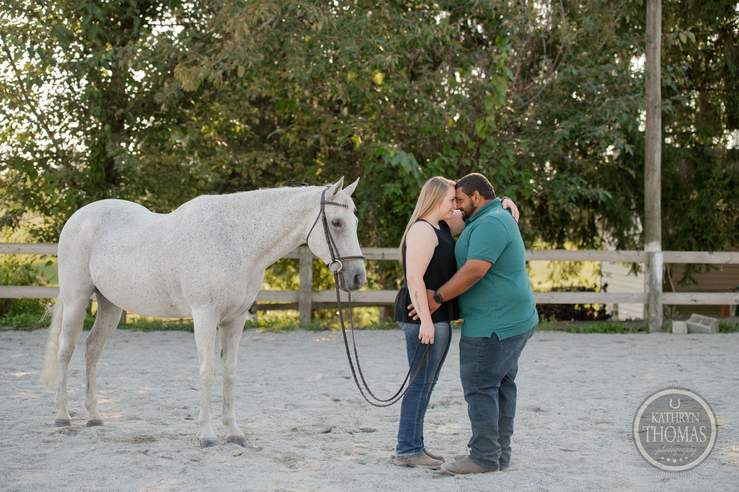 A white horse stands next to a couple sharing a tender moment in a paddock.