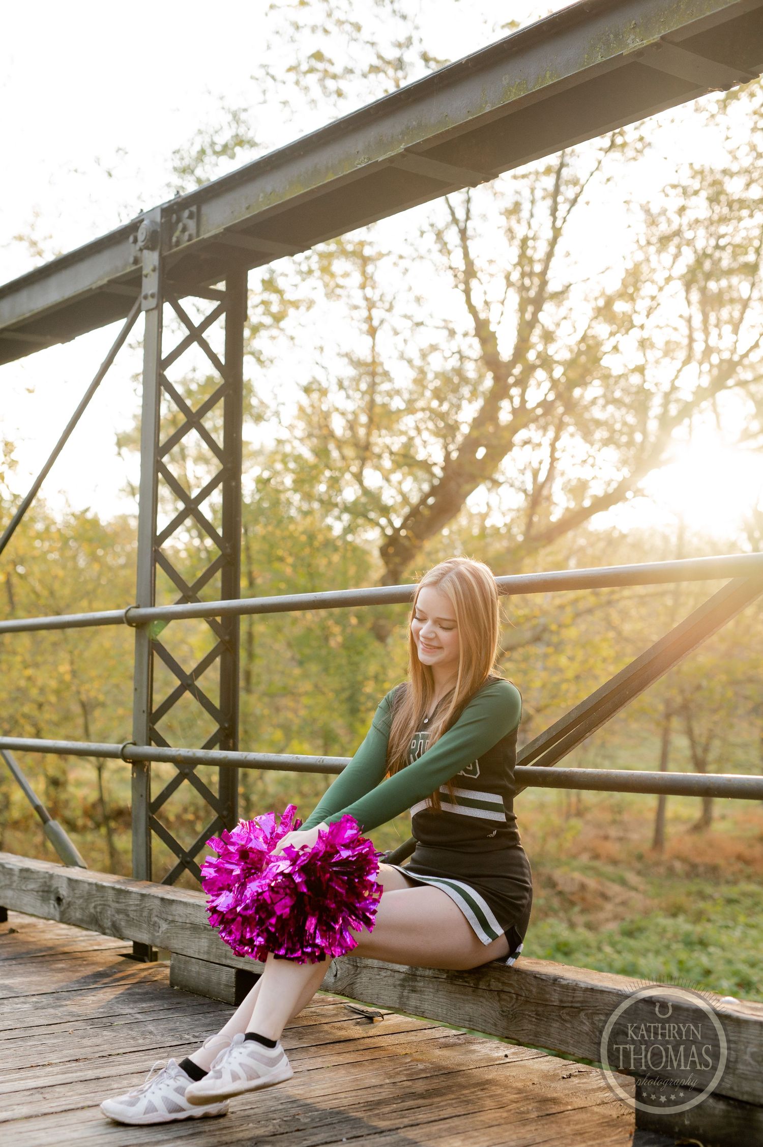 Cheerleader with bright pink pom poms sits on wooden bridge railing during golden hour sunset.
