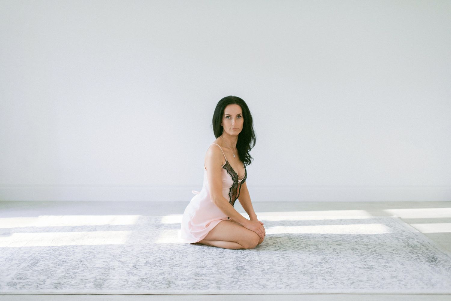 Someone in a light pink slip dress kneels gracefully on a white floor in a bright minimal setting.