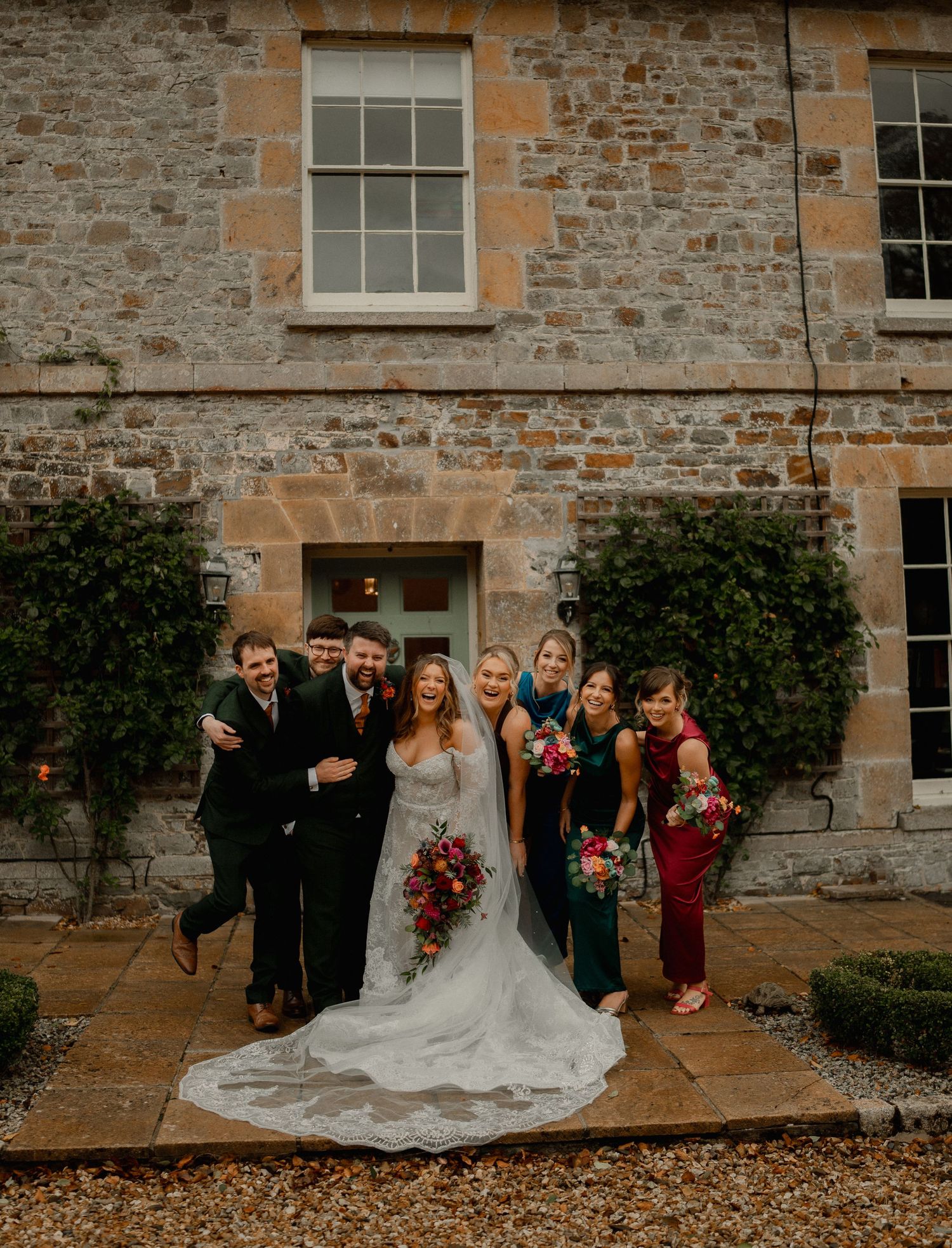 Bride in white gown poses with wedding party outside a stone manor house, holding vibrant red and pink bouquets.