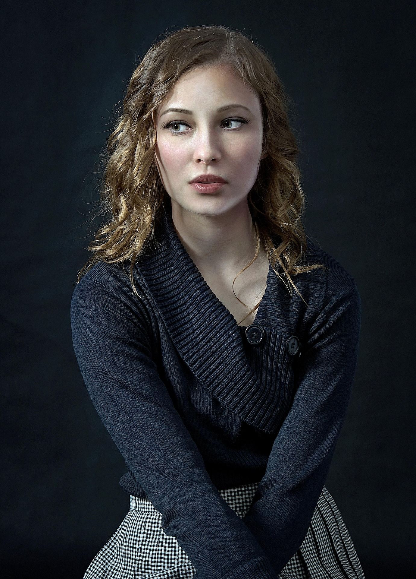 Jeune femme aux cheveux bruns bouclés, vêtue d'un cardigan en maille foncée et d'une jupe pied-de-poule, posant sur fond sombre