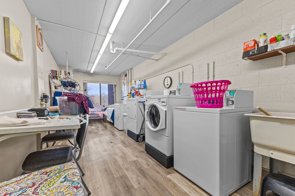 Community laundry room with multiple washers and dryers on wood-look flooring with storage shelves.