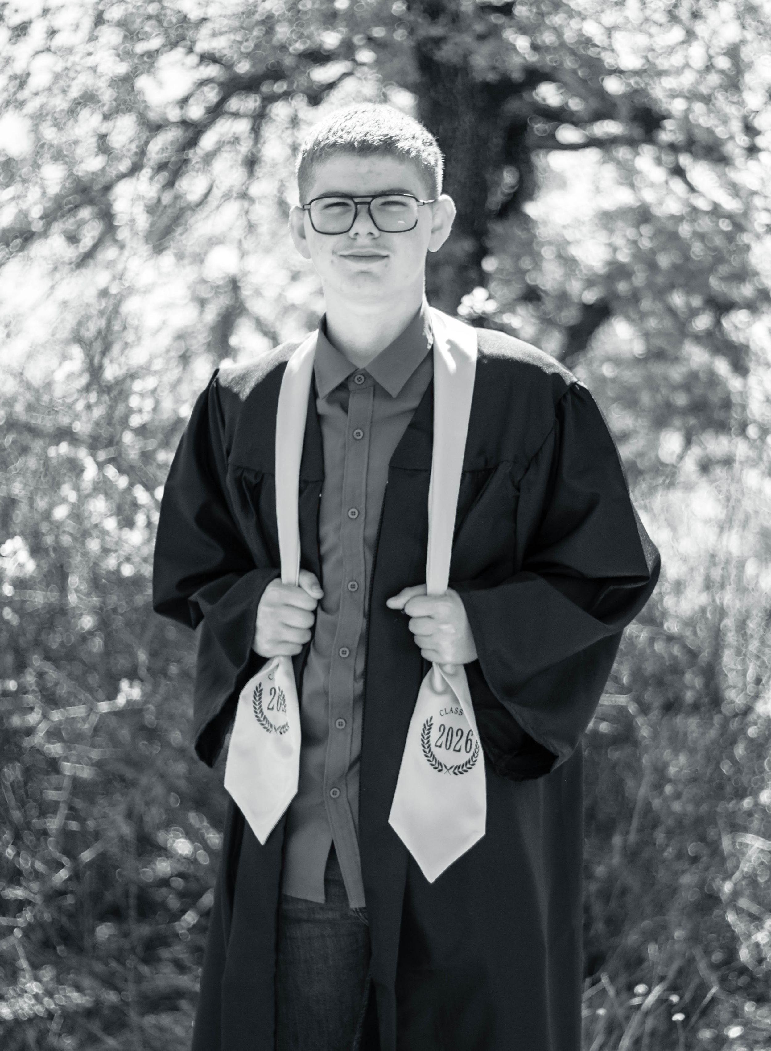 Young graduate in black gown and 2020 stoles poses outdoors in black and white photo.