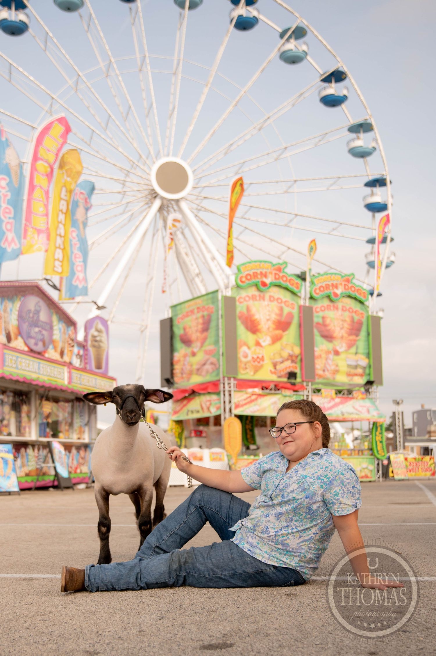 A young boy kneels beside a sheep at a county fair with a large Ferris wheel and colorful game booths in the background.