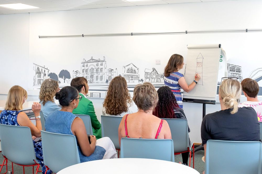 Students seated in blue chairs watch an instructor draw on a whiteboard with city skyline sketches during a classroom lesson.