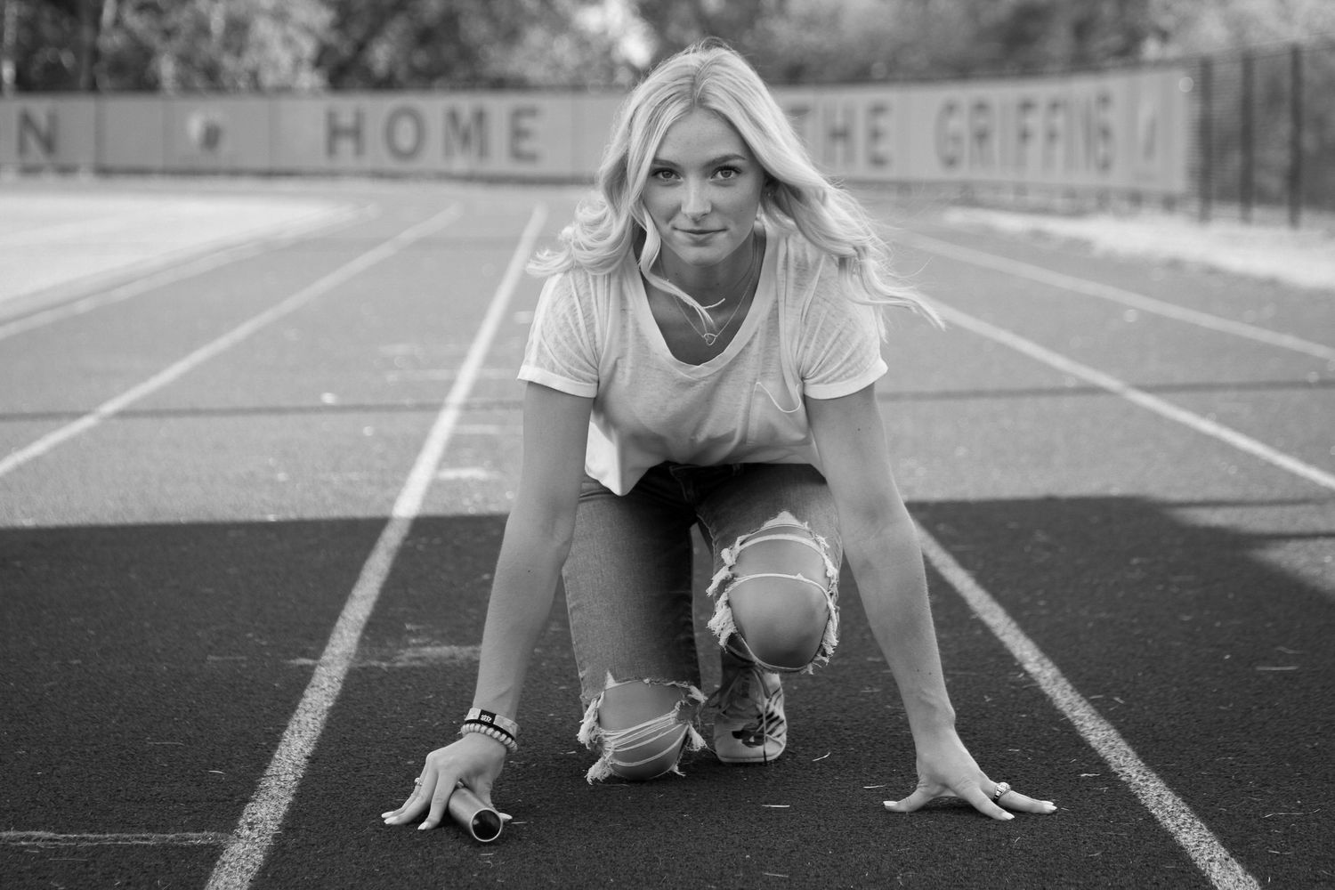 A young woman holding a baton crouches in the starting position on a high school track during her Kansas City senior photo session.