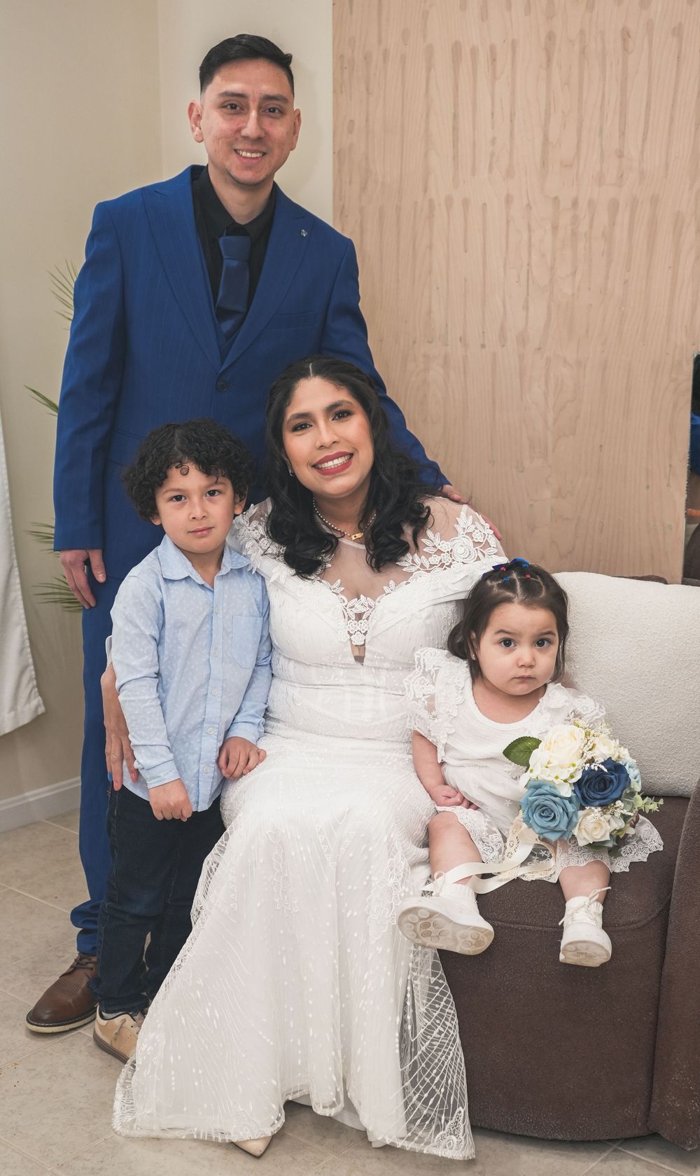 Family portrait at a wedding with groom in blue suit, bride in white lace gown, and two young children.