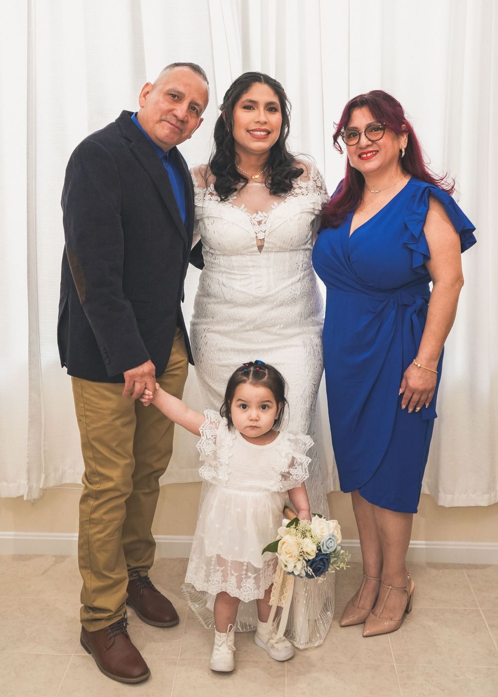 Bride in lace gown poses with family including man, woman, and young girl in white dress.