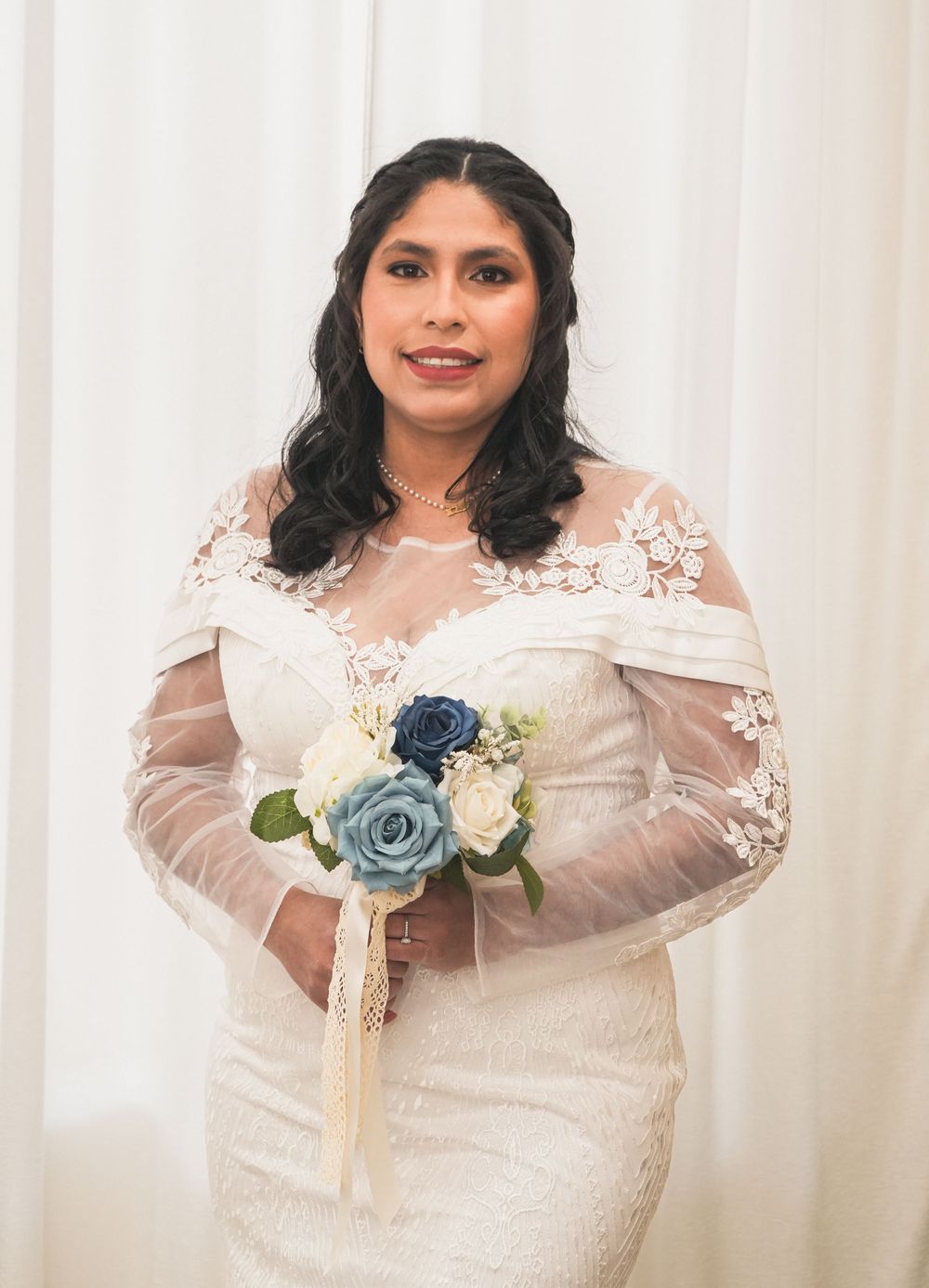 Bride in lace wedding gown with sheer sleeves holding blue and white floral bouquet.