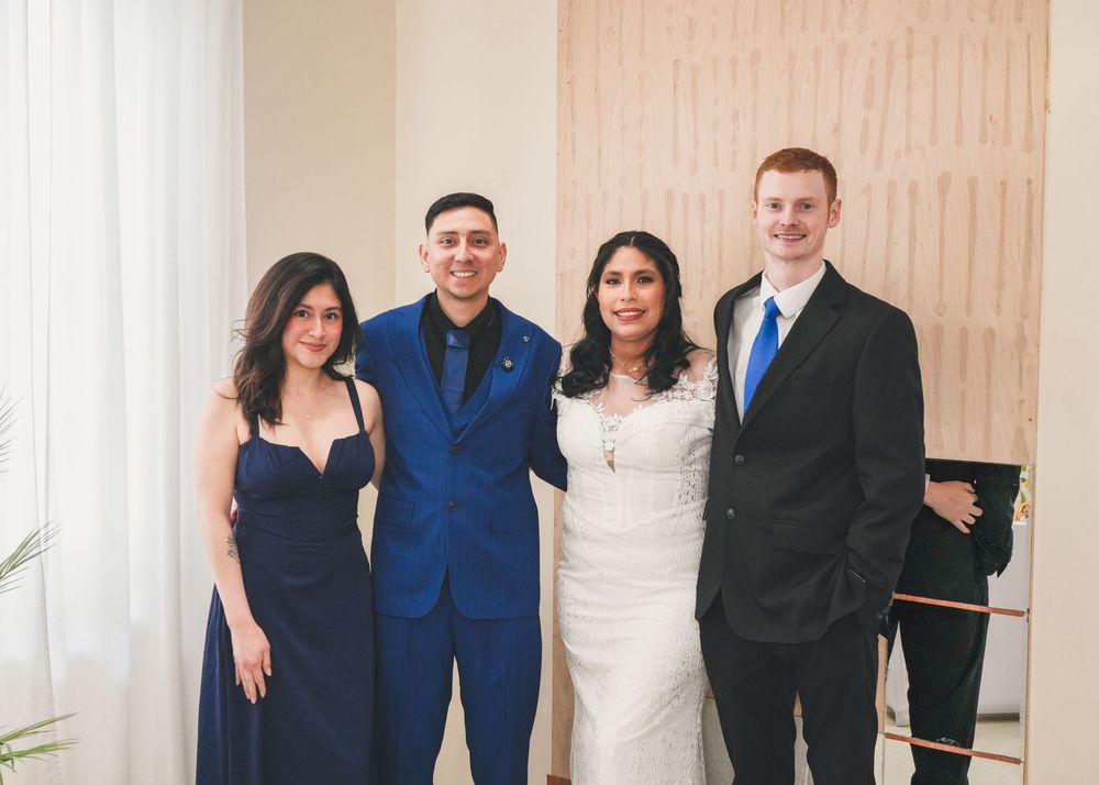 Four elegantly dressed guests pose together at a wedding ceremony, with the couple in wedding attire at center.