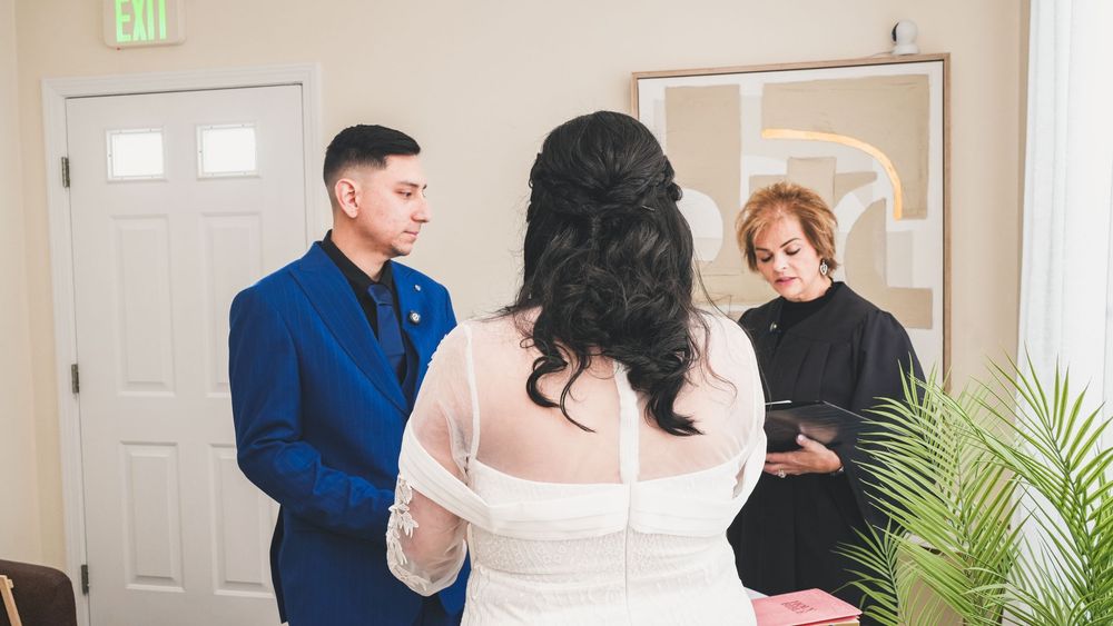 Couple exchanging wedding vows before an officiant in a bright indoor ceremony, groom in blue suit.