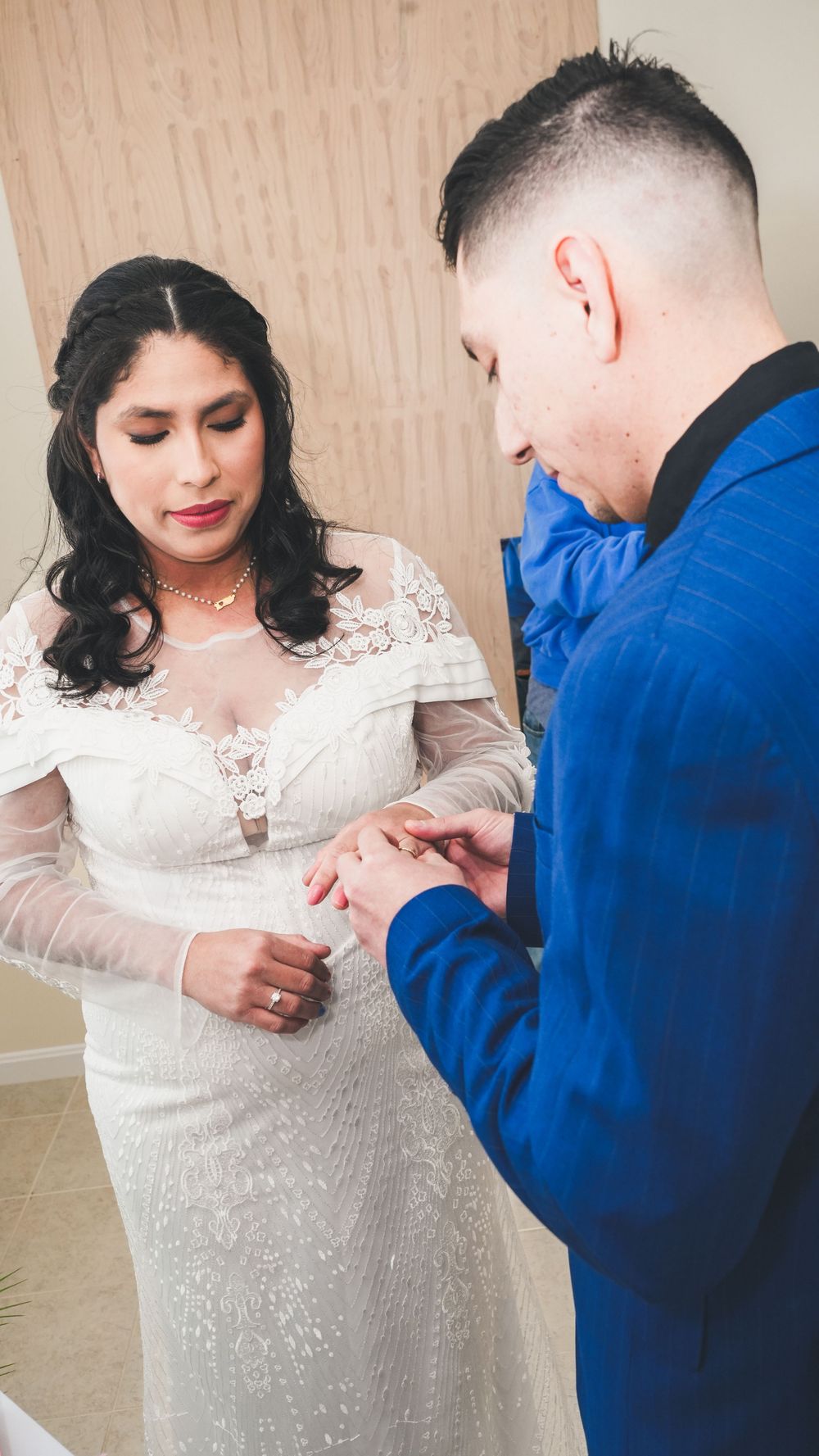 Groom in blue suit placing wedding ring on bride's finger during ceremony, bride wearing white lace gown.