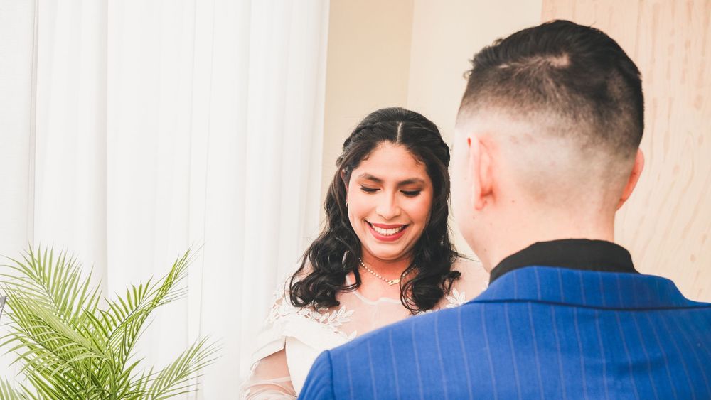 Smiling bride in pink dress faces groom in blue suit during intimate wedding ceremony indoors.