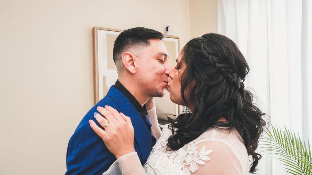 A couple shares a romantic first kiss at their wedding, bride in white lace gown and groom in blue suit.