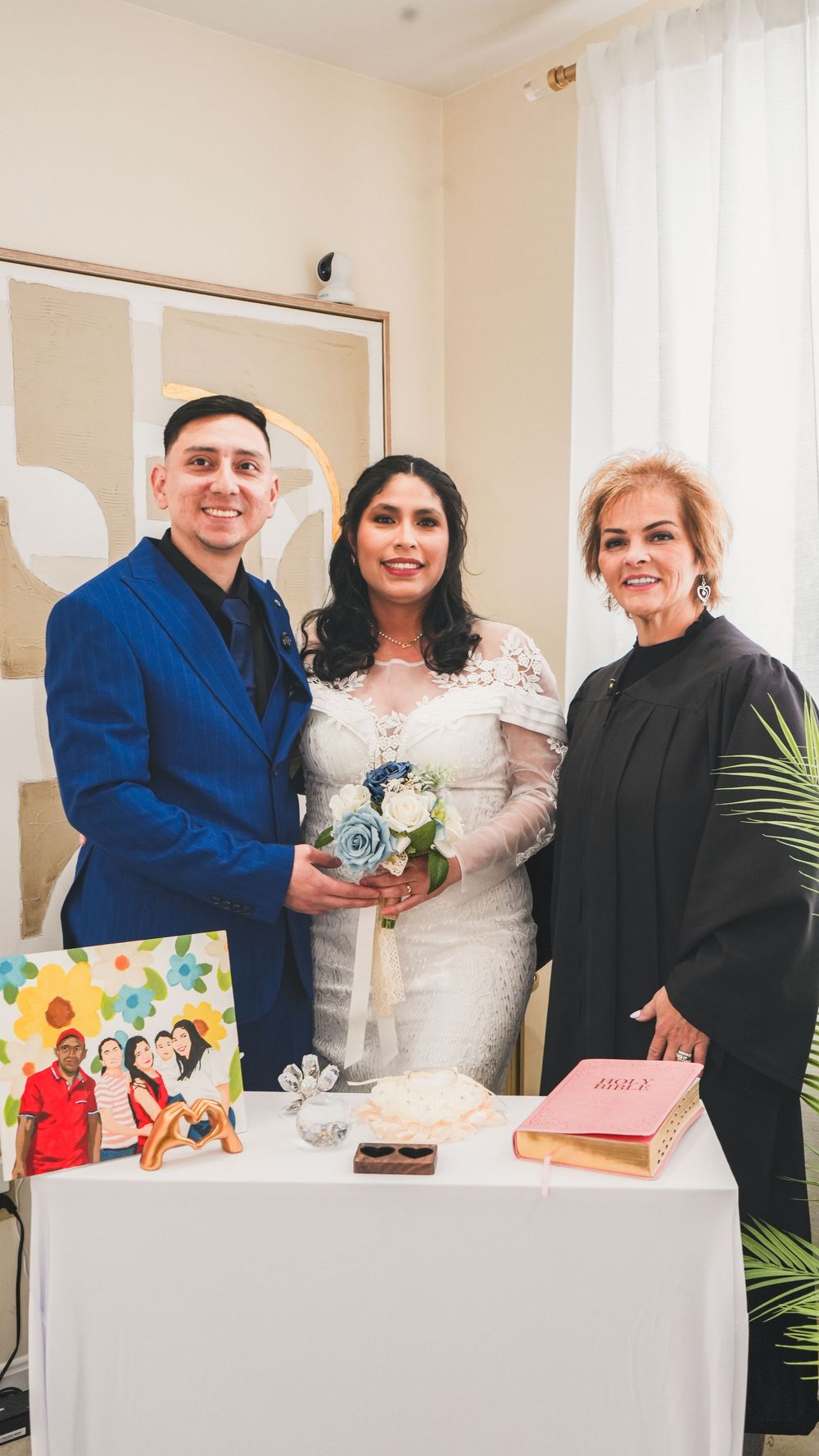 Bride and groom in blue suit pose with officiant at wedding ceremony altar with bouquet and Bible.