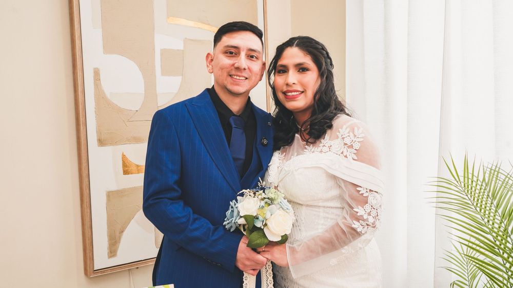 A couple poses together on their wedding day, the groom in a blue suit and bride in a white lace gown holding a bouquet.