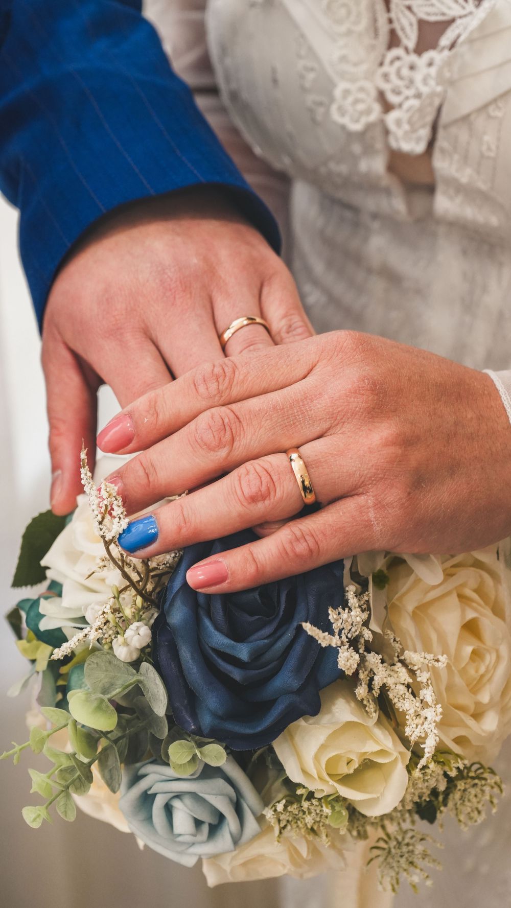 Newlyweds displaying gold wedding rings on hands resting over blue and white rose bridal bouquet.