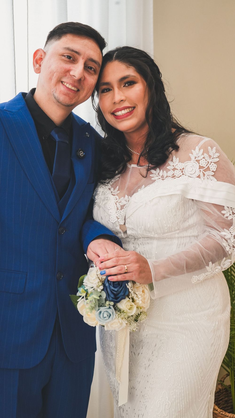 Happy newlywed couple posing together, groom in blue suit and bride in lace wedding dress with bouquet.