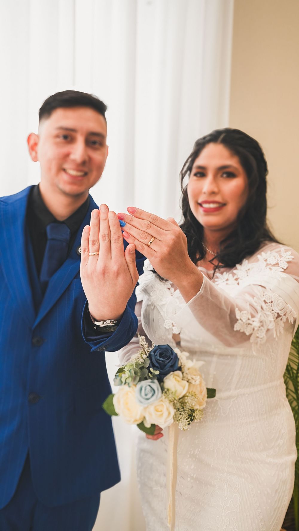 Smiling bride and groom showing off wedding rings at indoor ceremony, bride holding blue rose bouquet.