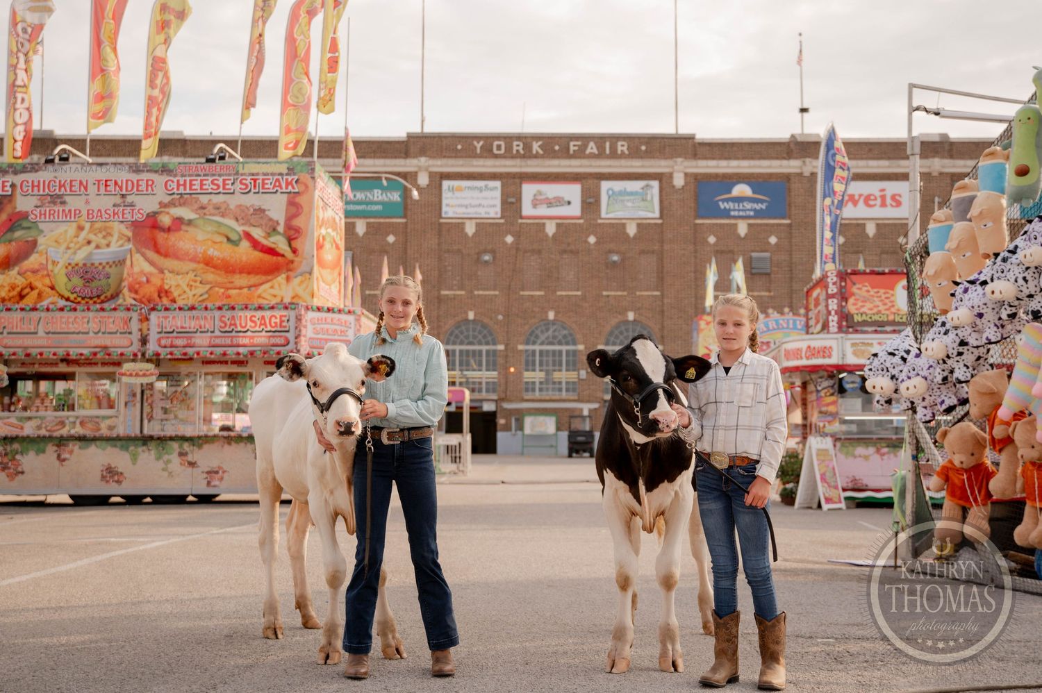 Two children hold calves at the York Fair, with colorful carnival games and food stands visible in the background.