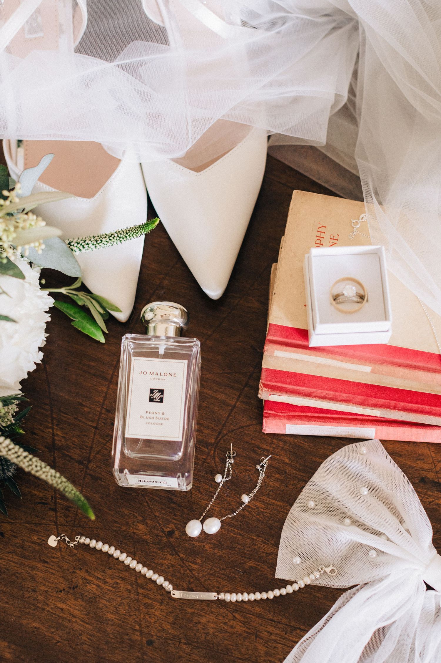 Bridal flat lay featuring Jo Malone perfume, white heels, pearl jewelry, wedding rings, red books, and floral bouquet on wood.
