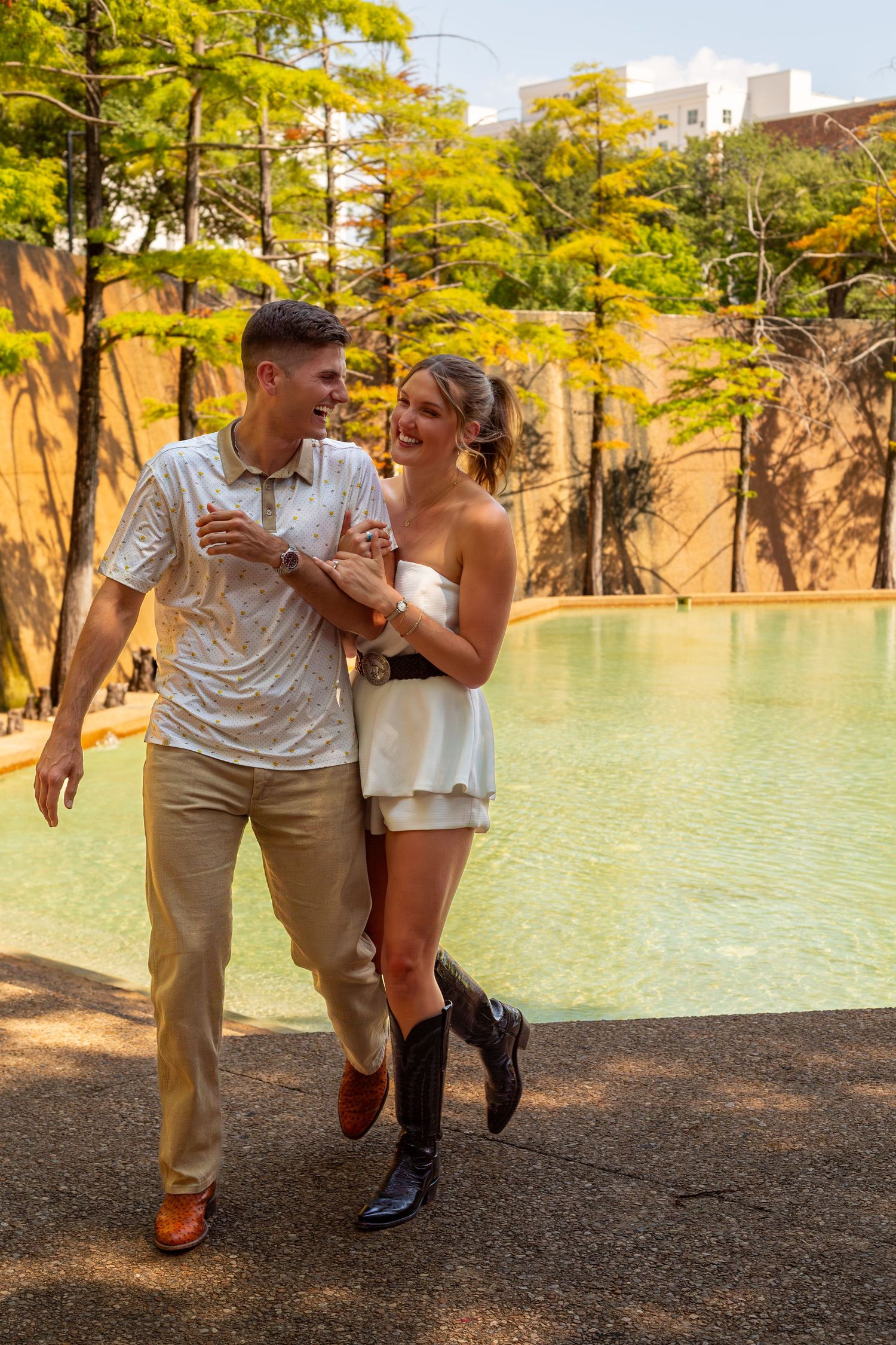 A smiling couple walks hand-in-hand by a turquoise pool, woman in white dress and boots, man in polo and khakis.