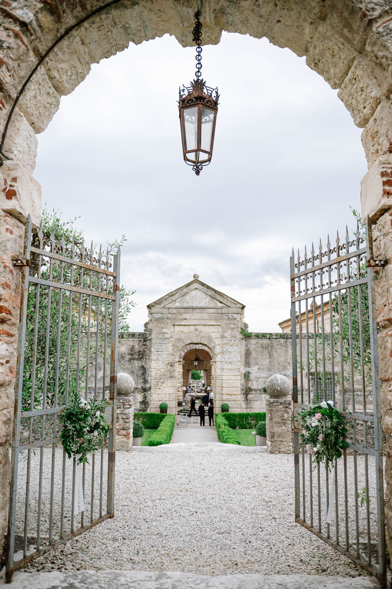 Elegant iron gates open to a gravel path leading to a stone archway at a historic European villa wedding venue.