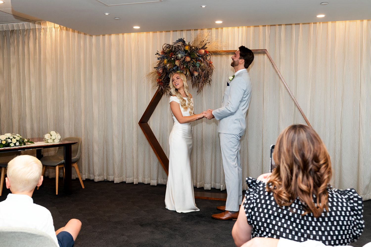 Couple exchanging vows at indoor wedding ceremony beneath geometric arch with floral arrangement.