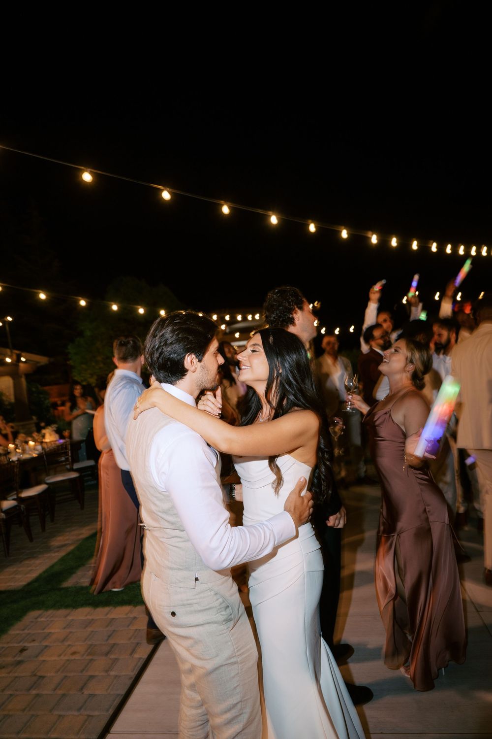 Couple sharing a romantic kiss on outdoor wedding dance floor at night with string lights and guests celebrating with glow sticks.