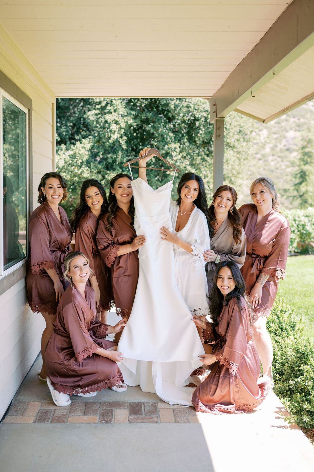 Bride holds wedding dress surrounded by bridesmaids in mauve robes on a porch.
