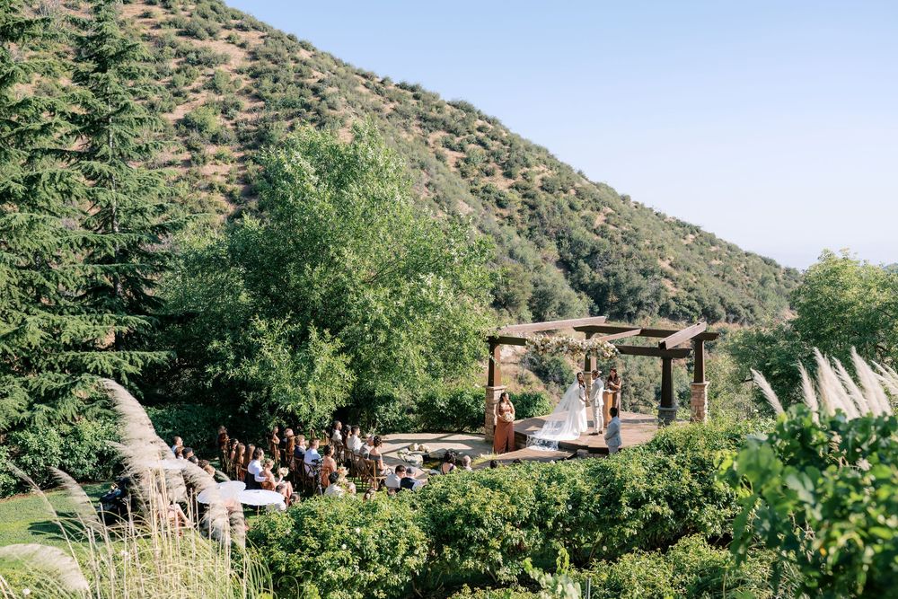 Outdoor wedding ceremony set against a lush green hillside with guests seated beneath a wooden pergola altar.