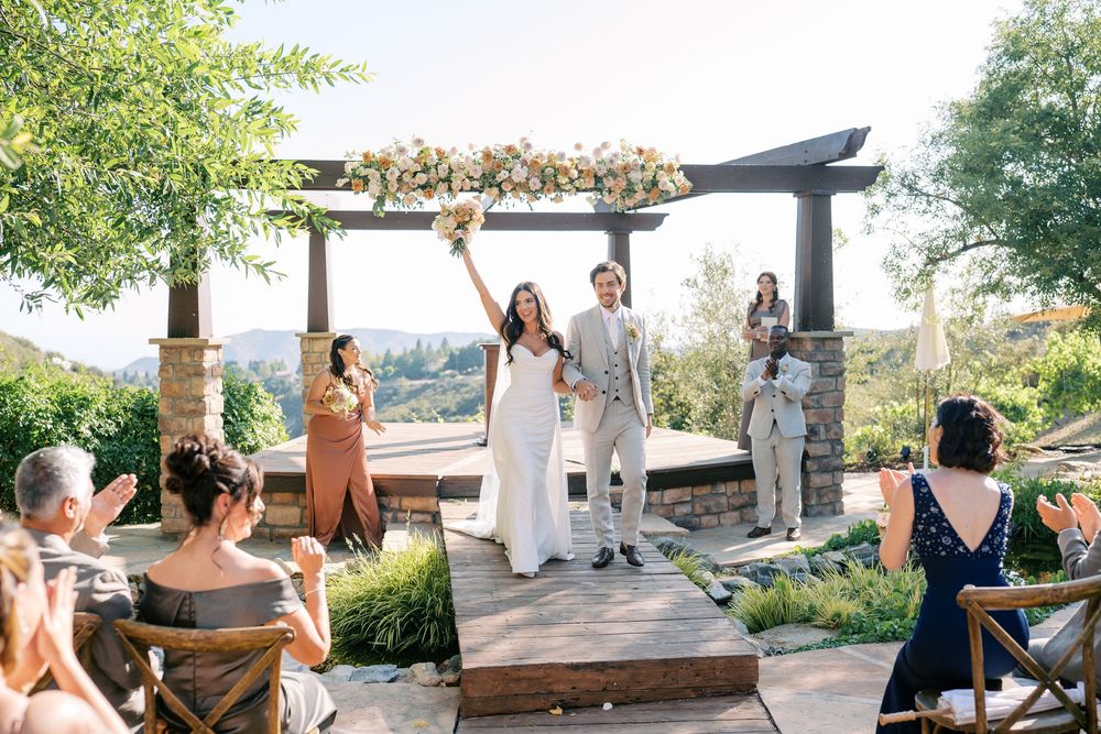 Bride and groom raise hands in celebration at outdoor wedding ceremony under floral arch with scenic hillside views.